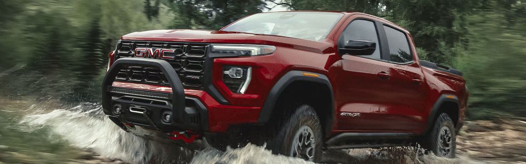 A Red GMC Canyon AT4X splashes through a shallow stream on a forest trail, kicking up water as it drives.
